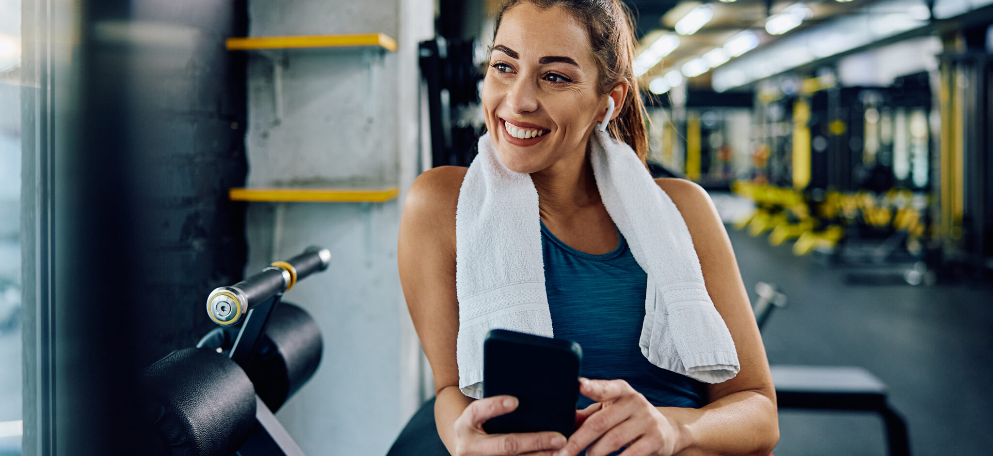 Happy sportswoman using smart phone while listening music over earbuds in health club and looking away. Copy space.